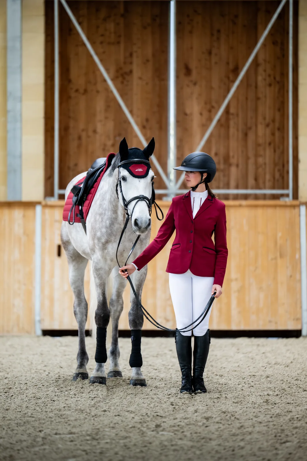 Rider in the red Equiline EQCAT show jacket next to a grey horse wearing the red EQCRISCE saddle pad and ear bonnet in an indoor arena.