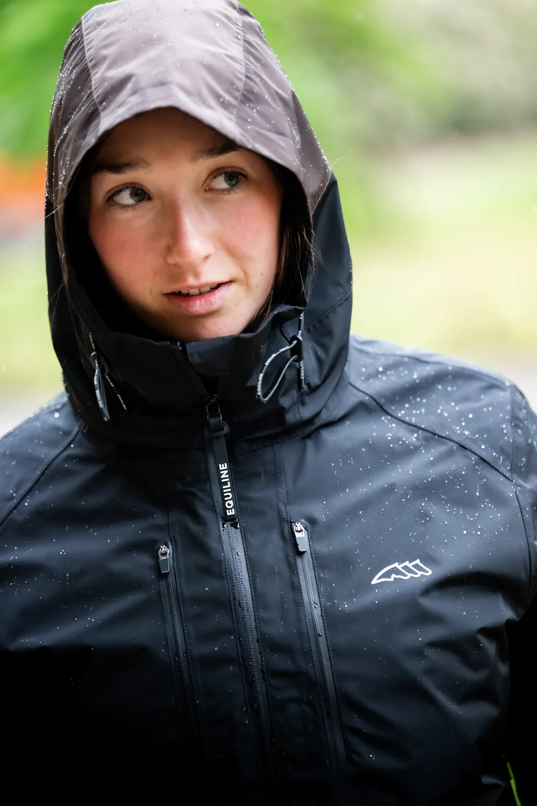 Woman wearing the hood of the black Equiline EQCROOF waterproof jacket in the rain.
