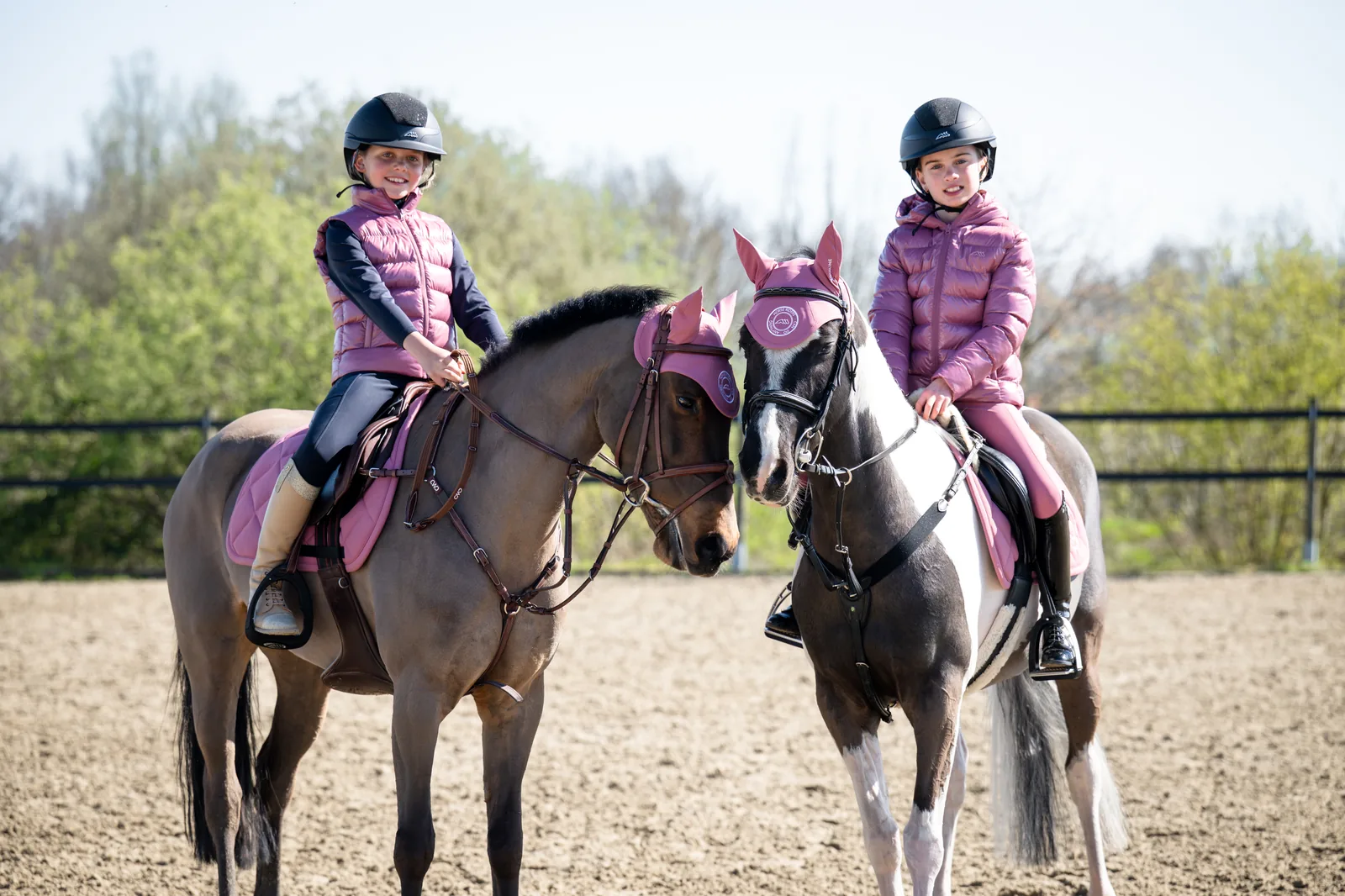 Two girls riding ponies, one in a pink Equiline gilet and the other in a pink puffer; the ponies wear the pink EQPERRY saddle pad and ear bonnet.