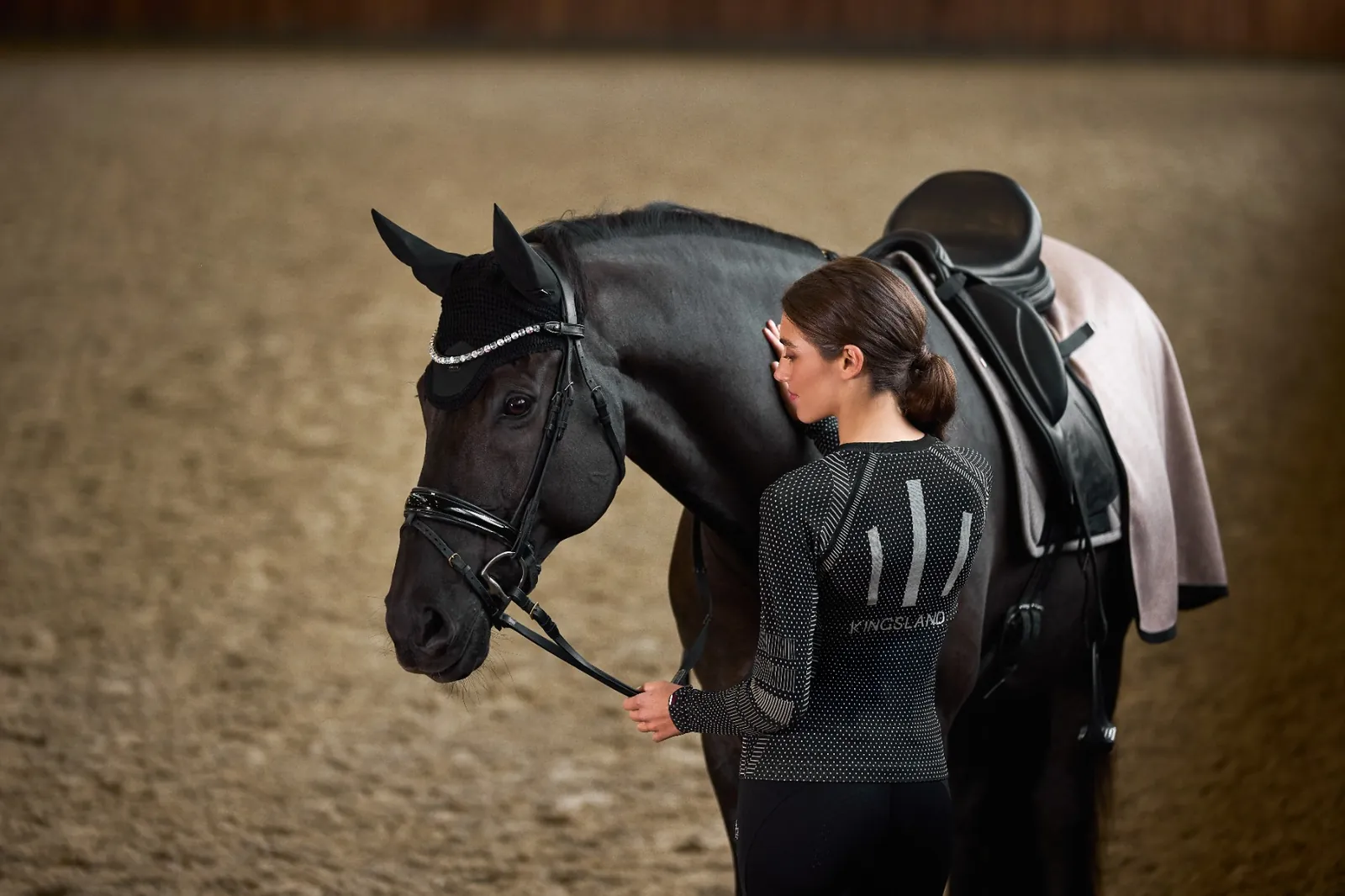 Cavalière de dos avec le t-shirt noir sans coutures Kingsland Alicia à côté d'un cheval noir sellé, dans un manège.