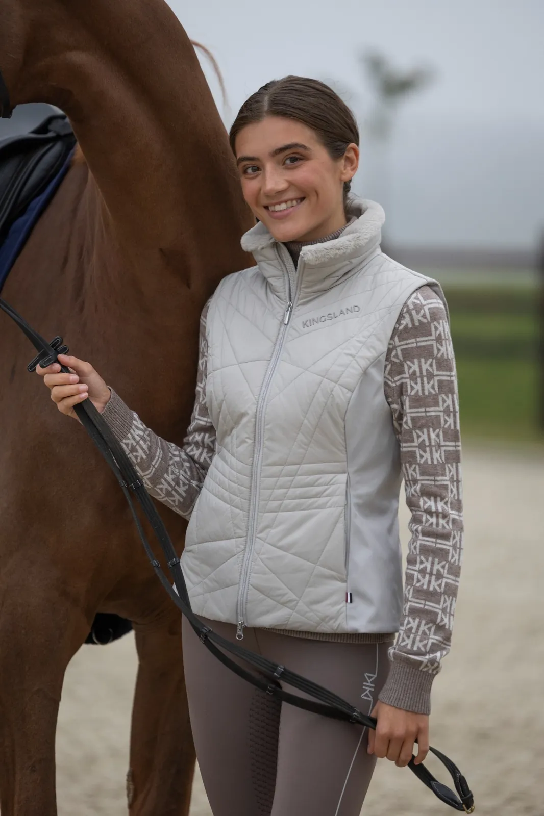 Cavalière souriante en legging d'équitation Kingsland Amy marron à côté de son cheval alezan, dans une carrière brumeuse.