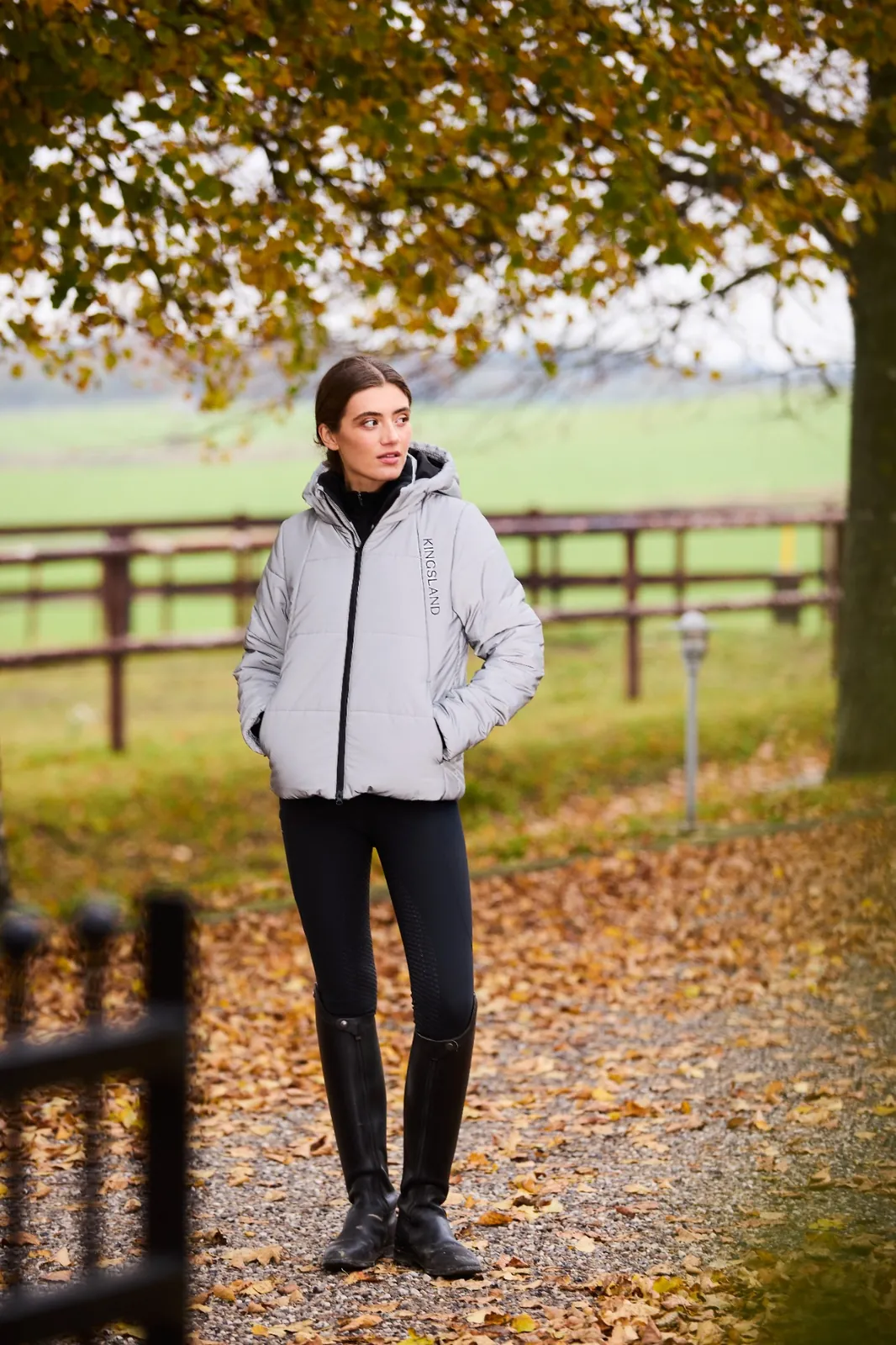Femme portant le manteau matelassé réfléchissant argent Kingsland Aurora, legging noir et bottes d'équitation, sur un chemin couvert de feuilles d'automne.
