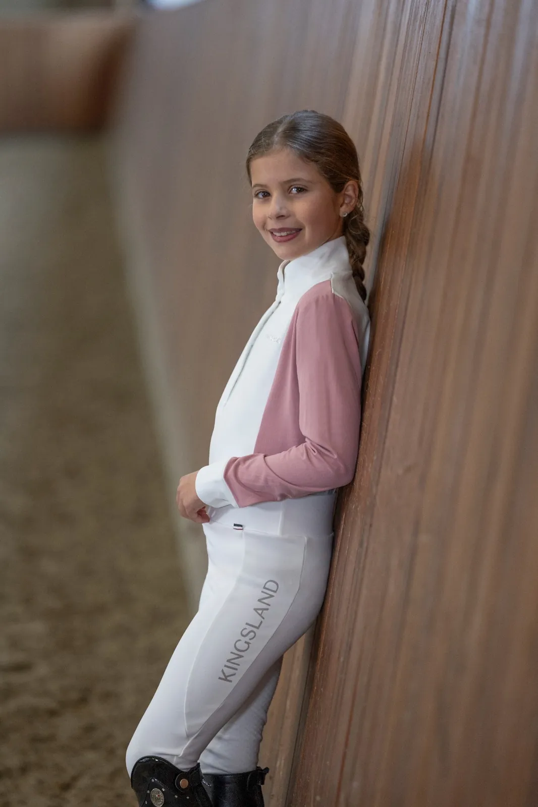 Fillette souriante appuyée contre le mur en bois du manège, avec le polo de concours Kingsland Adora blanc à manches roses et le legging blanc Kingsland Alexis.