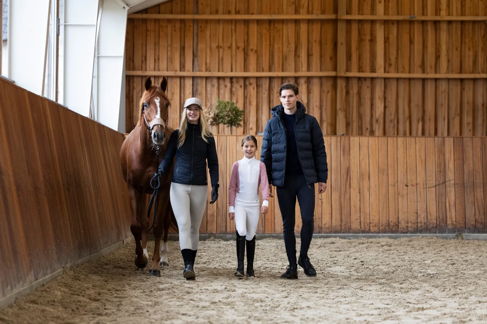 Une cavalière avec son cheval alezan, une fillette en polo de concours et un homme marchent ensemble dans un manège en bois.