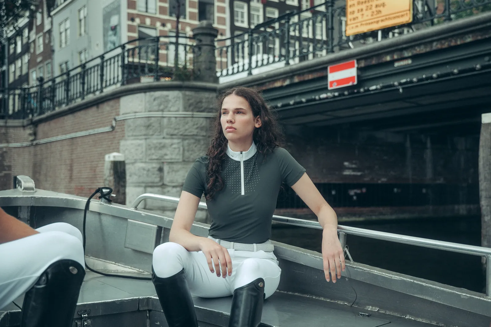 Rider sitting on a boat on an Amsterdam canal wearing the RG Deep Forest green short-sleeved show polo and white high-waisted riding breeches, with tall black boots.