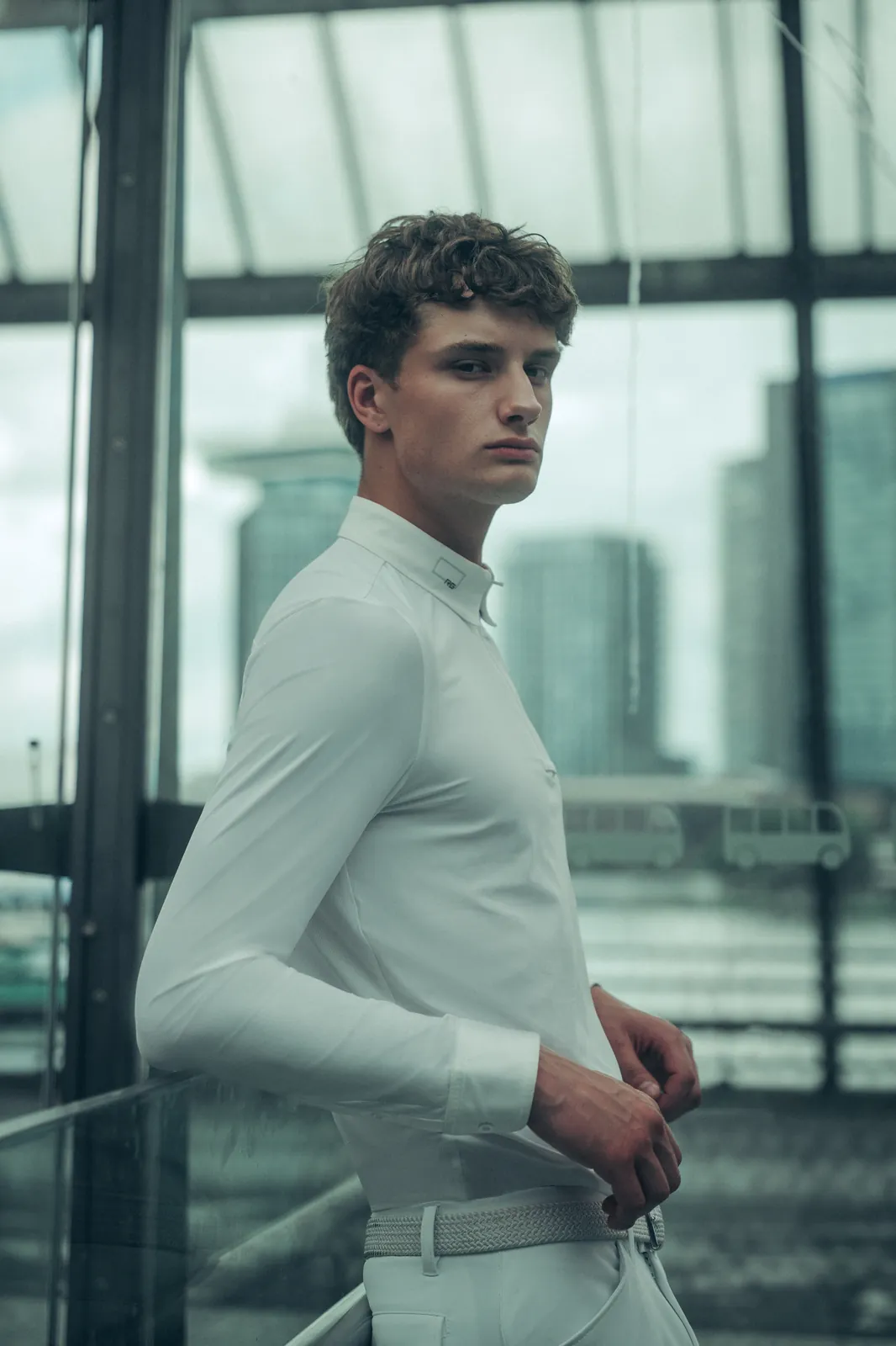 Male rider leaning on the railing of a glass-walled lookout in Amsterdam, wearing the RG white long-sleeved men's show polo and white breeches.