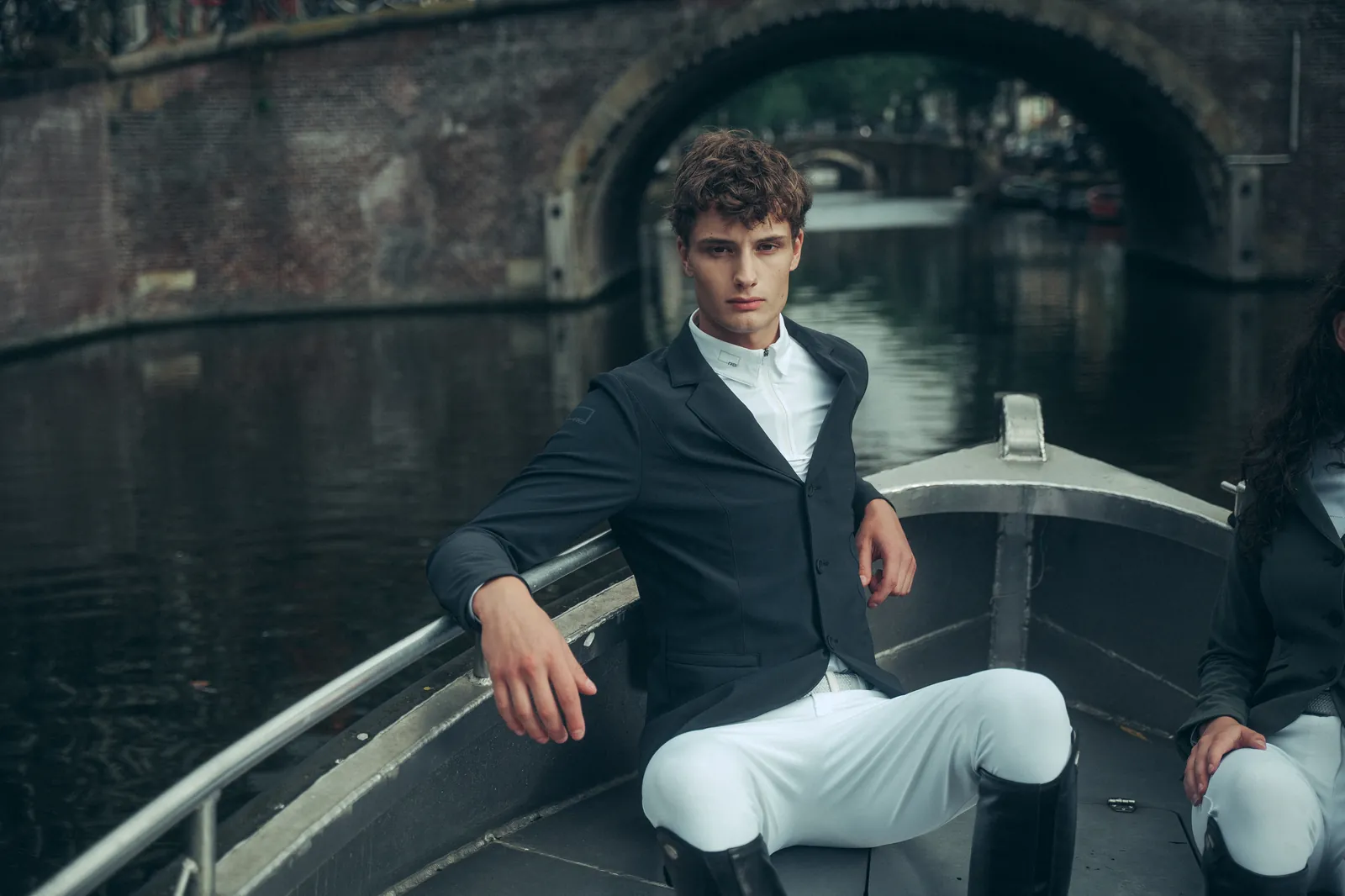 Male rider sitting at the bow of a boat under an Amsterdam bridge, wearing the RG navy competition jacket, a white show polo and white breeches.